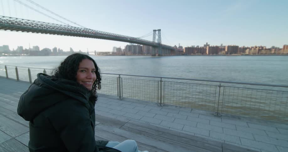 A woman sitting looking at the water and a city bridge turns her head and smiles