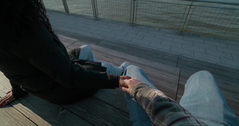 Point of view footage of a young couple sitting on a bench in front of the river close to the city skyline holding hands