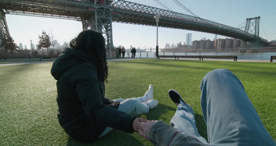 A man lies on the grass in front of the water with the city skyline in the distance and holds a womans hand as she sits next to him