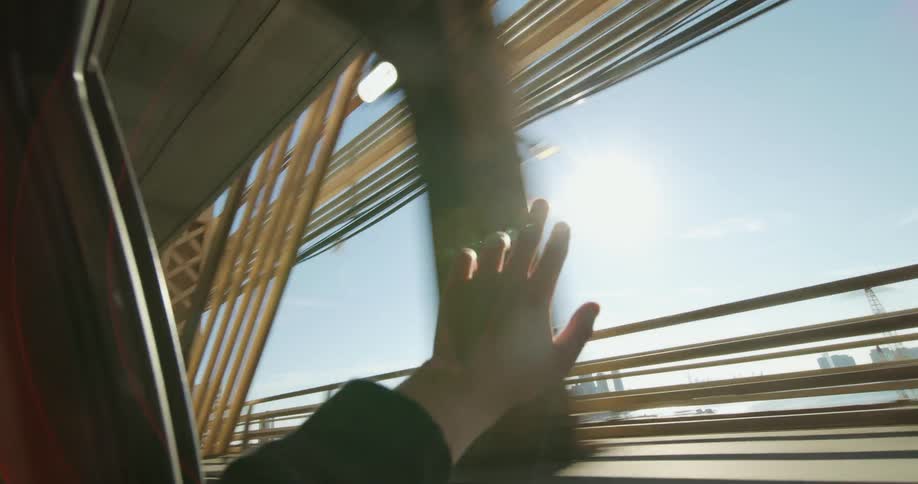 A hand reaching out of the back seat passenger window as a car travels over a city bridge on a bright sunny day