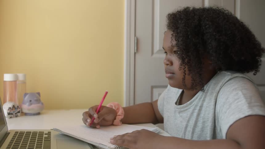 Young girl studying on her room while she looks at her laptop and write down on her notebook