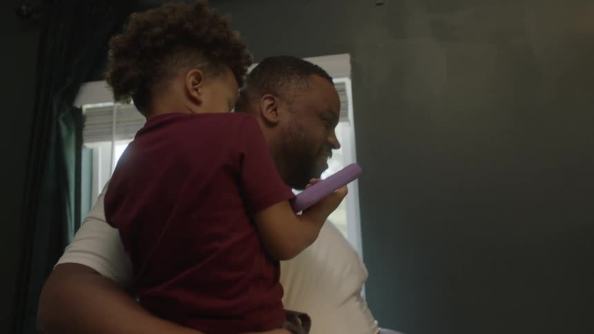 A man carries a small child with his arm while participating in a video conference using a laptop on a nearby desk
