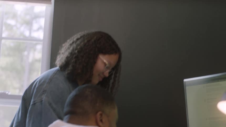 A man is sitting at a desk when a girl brings a notebook and laptop over to him