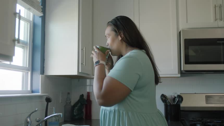 A woman walks through her kitchen and goes to the window and looks out as she takes a drink of her coffee
