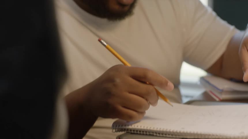 Man sitting at a desk at home reading from a laptop taking notes on a notepad with a pencil