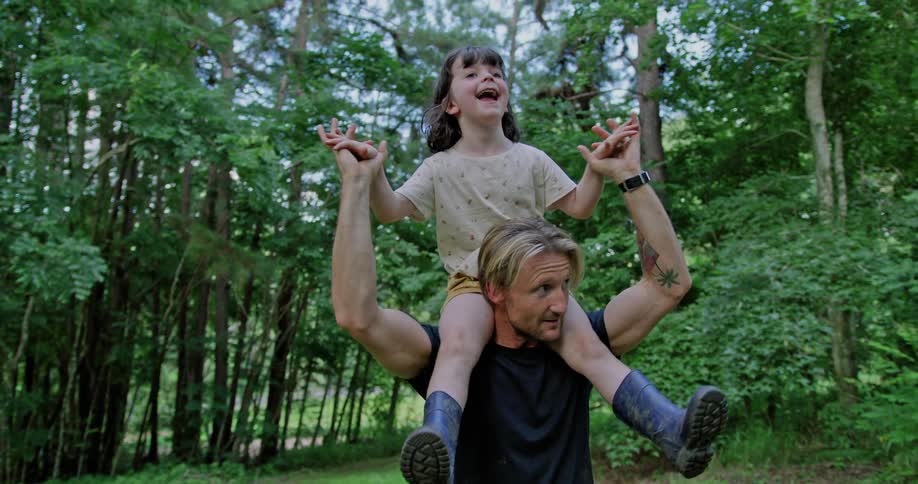 A girls smiles as her dad carries her on his shoulders in a forest