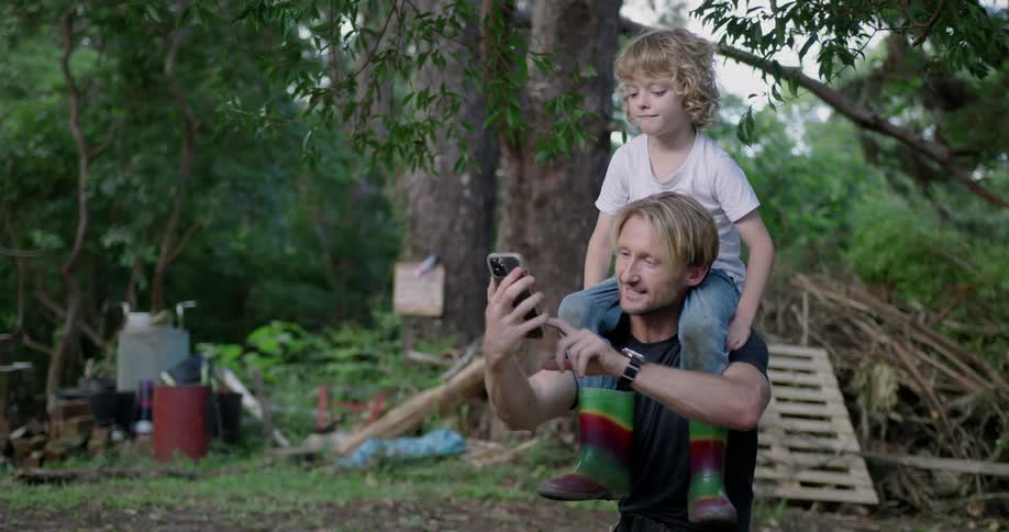 A girl sits on her dads shoulders and they both smile as he holds up his cell phone and takes a photo of them