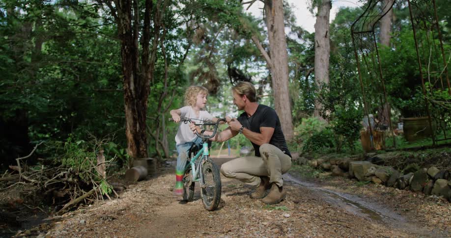 A little boy rides by as a man is helping another boy on his bicycle