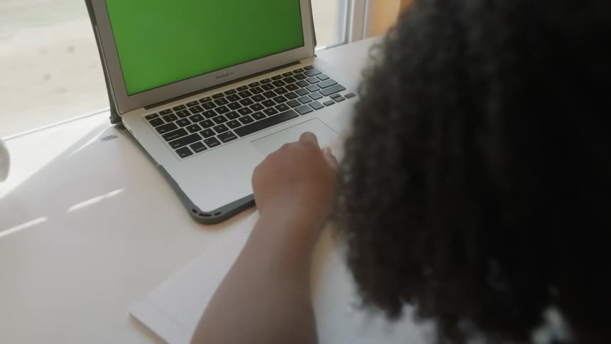 Young girl sitting at a desk in her bedroom in front of a laptop with green replaceable screen doing homework with pen and paper