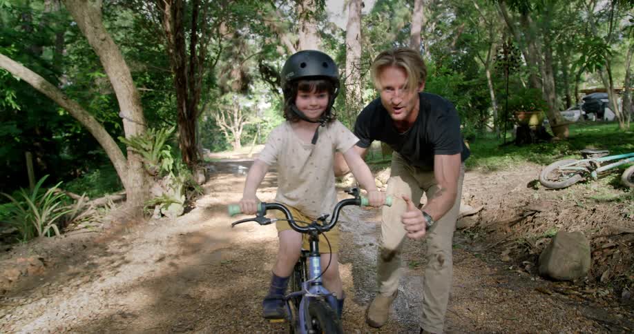 Dad celebrates with his arms in the air as his daughter rides her bike on the dirt path