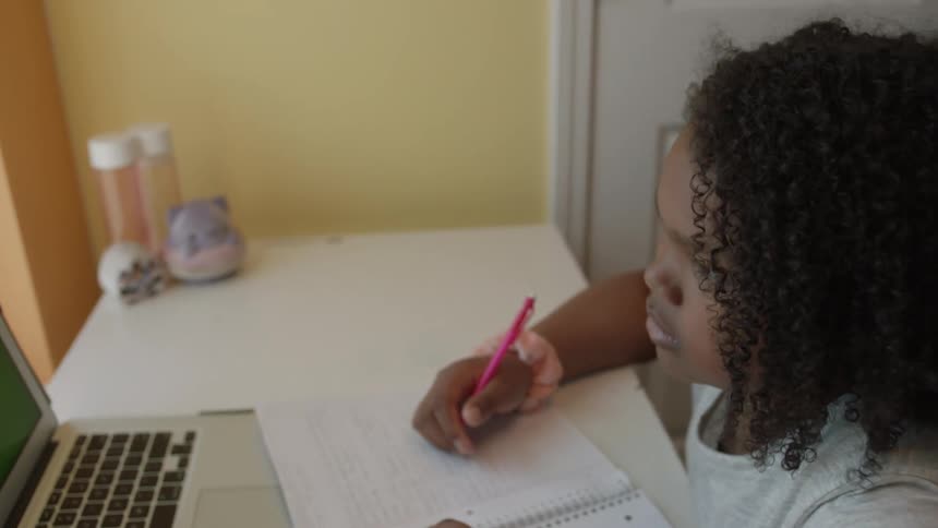 A girl looks at the screen of a laptop while writing in a notebook as she sits at a desk indoors