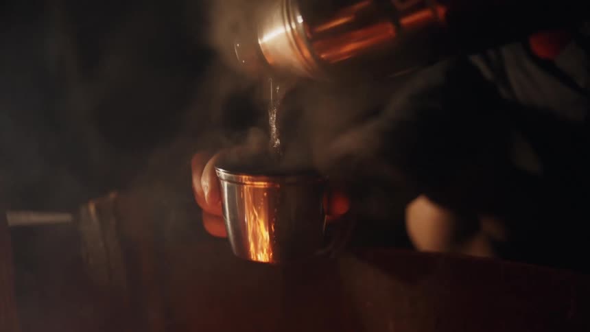 Person with black jacket puts water into cup from thermal flask next to campfire at night