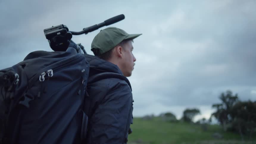 Man wearing backpack hikes across field under overcast sky