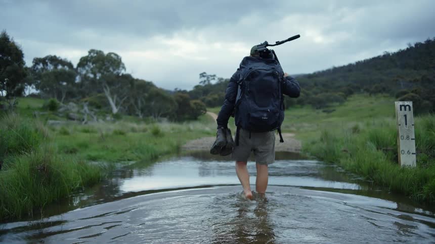 Man wearing backpack crosses water carrying boots