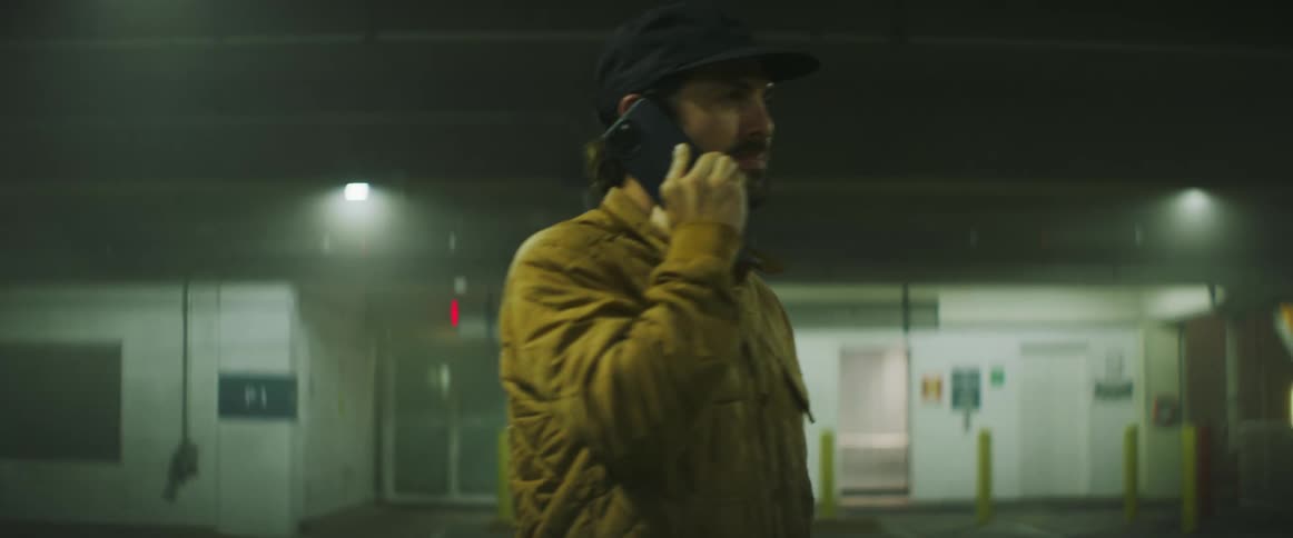Man wearing quilted jacket and baseball cap makes call on mobile phone while standing in parking garage