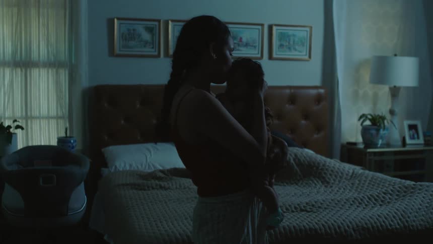 Mother kisses her daughter on her arms in front of the bed inside a dimly lit room at home