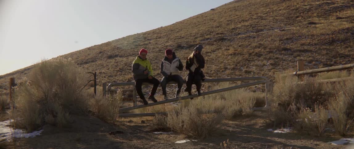 Three people sitting on fence by hill with relaxed thoughtful conversation vibe.
