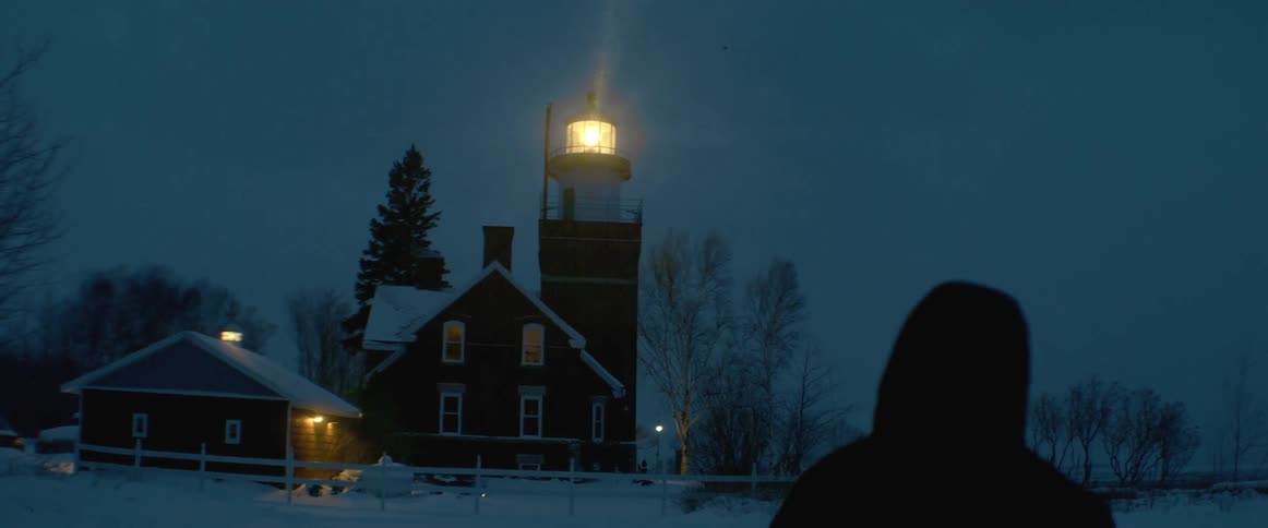 A person walking towards a house with a lighthouse in the middle of a rural area while its snowing