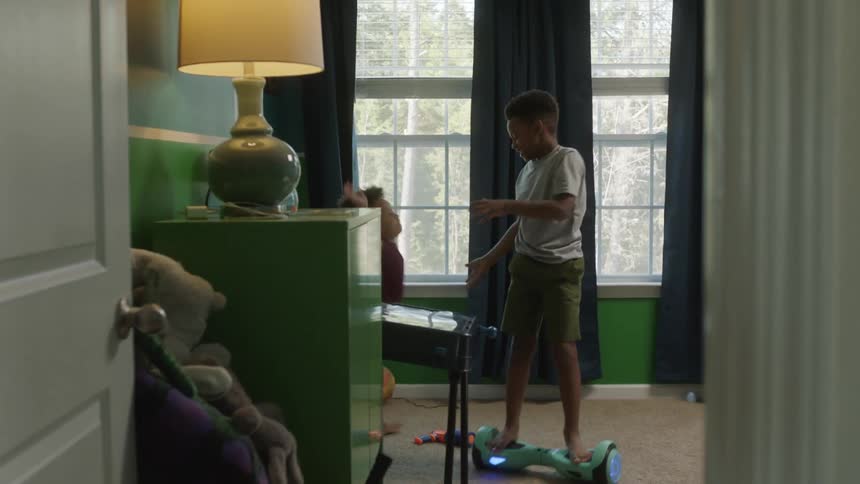 Two boys ride hoverboard next to windows and black curtains in room