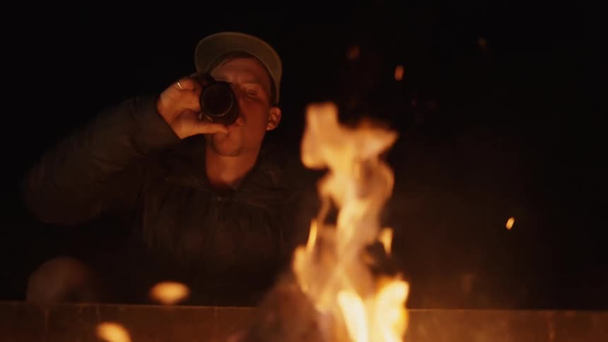 White man sitting next to a bonfire drinking a beer