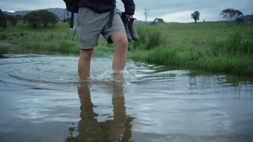 Man wades through water near river bank carrying boots