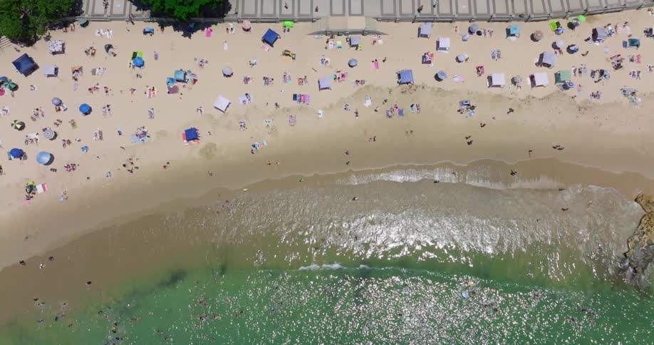 An aerial view of waves rolling in to beach with umbrellas and people filling the sandy shore up