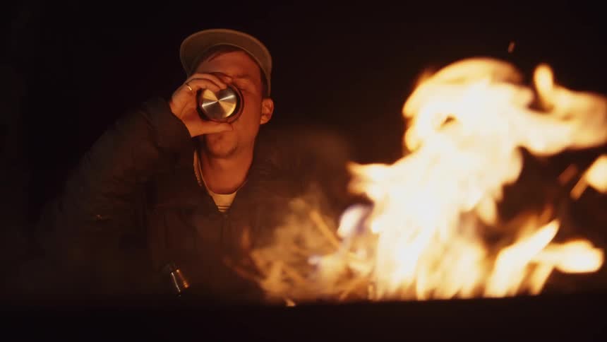 A man is deep in thought as he takes a drink and stares at a bonfire