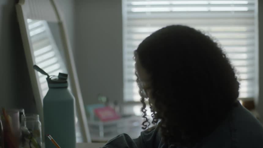 A young woman writes in a notebook while glancing up at her laptop as she sits at a desk indoors