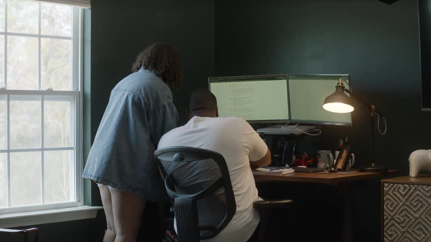 Woman walks into room to consult with man sitting at desk with dual monitor computer