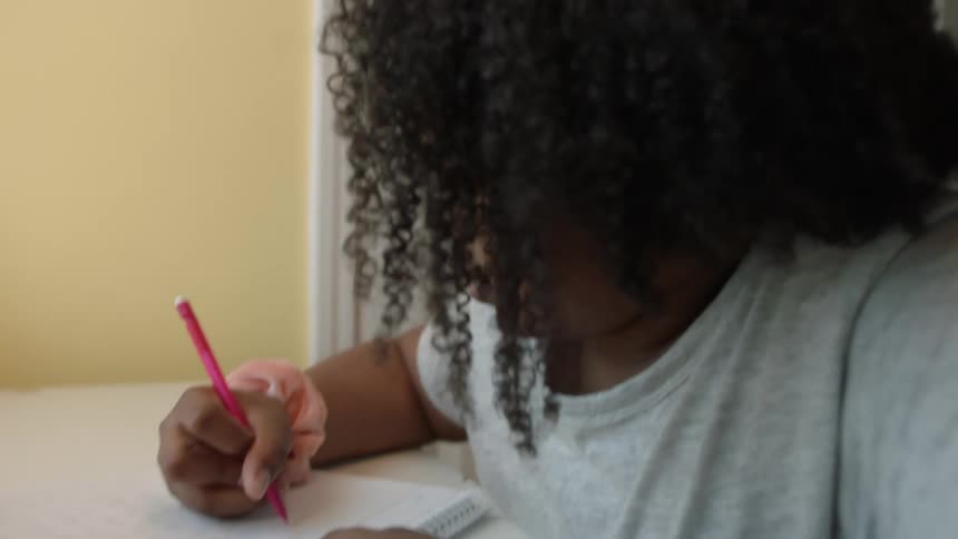 A girl writes in a notebook while looking up at the screen of a laptop as she sits at a desk