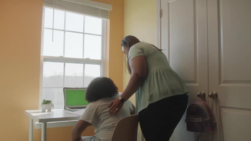 Mother checks on daughter sitting at table in front of laptop