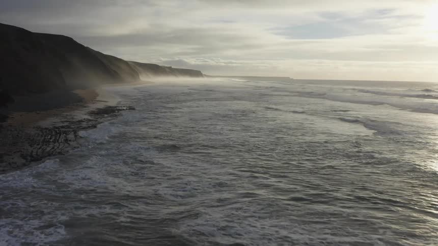 Ocean waves slowly roll into a beach below a cliff on a cloudy day