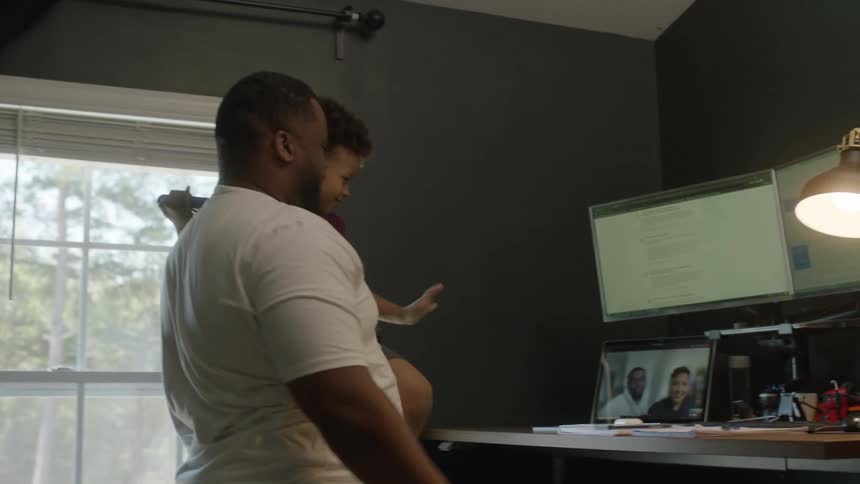 A man picks up his toddler to wave to the people on a video call on a tablet sitting on a desk