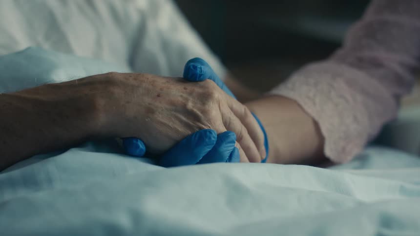 Visitor wearing glove holds hands with patient lying in bed