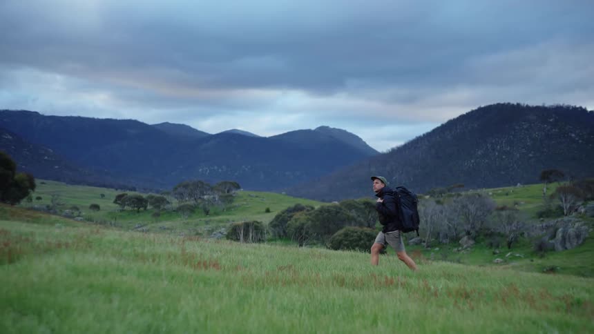 A man wearing shorts and a big backpack is walking across the grass with mountains in the distance