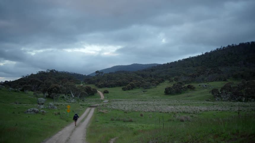A man wearing shorts and a big backpack is walking on a dirt road with mountains in the distance