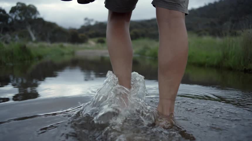 Man wades barefoot through water covering trail near forest covered slope