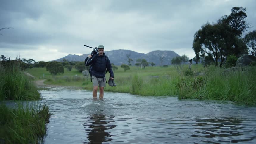 A man wearing shorts is walking through a stream carrying his boots and holding something over his shoulder