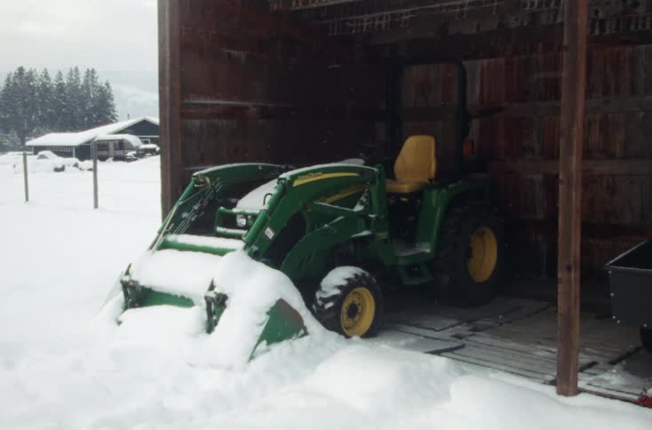 Snow covers farmhouse and exposed parts of farm vehicle that sits in shed