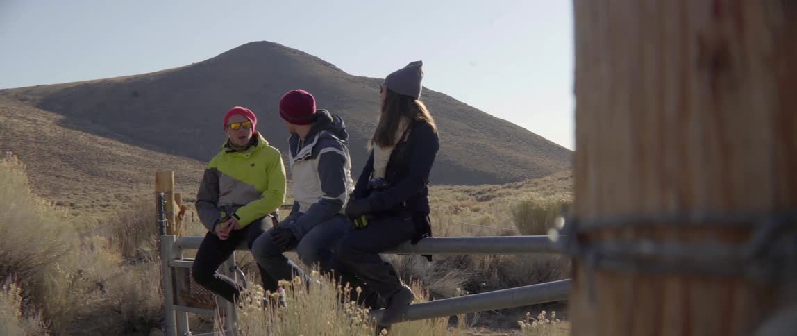 Three people sitting on fence by field talking with relaxed conversation vibe.