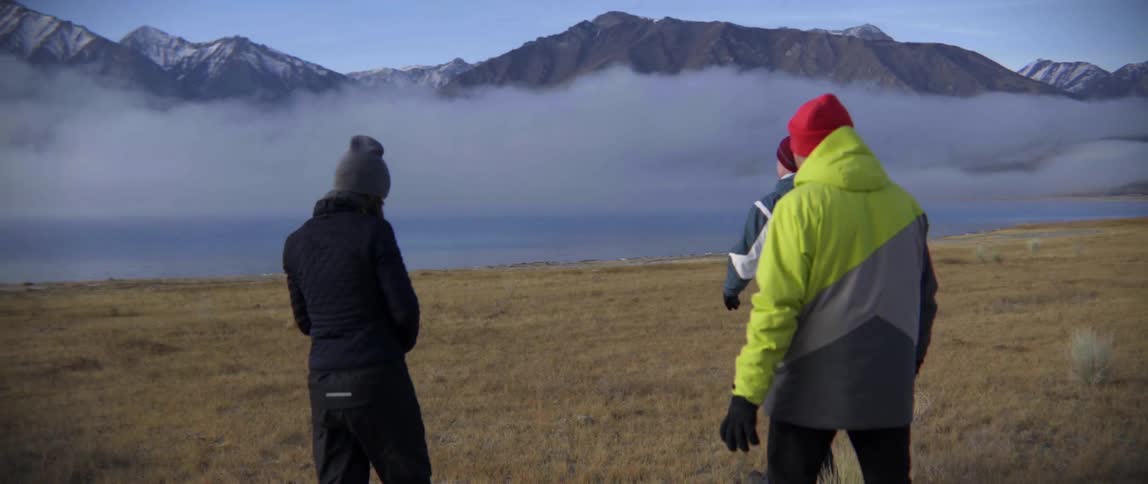 People walk along the grass towards a pond by a mountain range