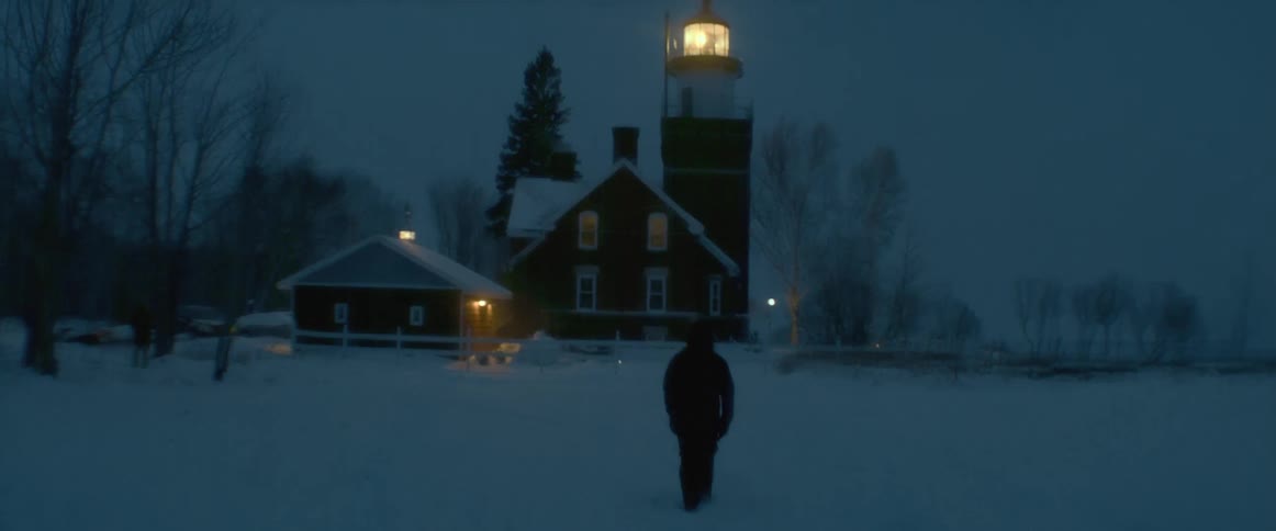 A bundled up person walks through heavy snow toward a house below a lighthouse