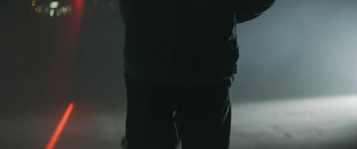 Man carries a skateboard on a dimly lit room filled with smoke near a red laser line on the floor
