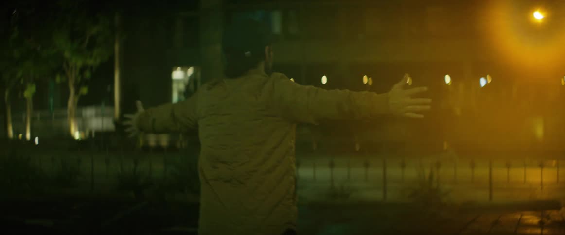 Man wearing jacket and baseball cap walks into street and spins with his arms wide as traffic moves along street