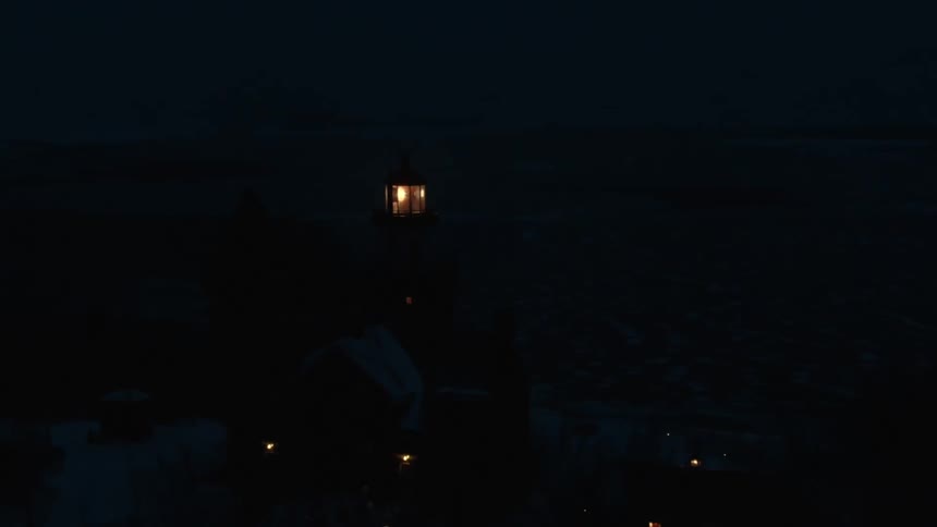 A beacon spins atop a lighthouse on a cliff on the coast at night