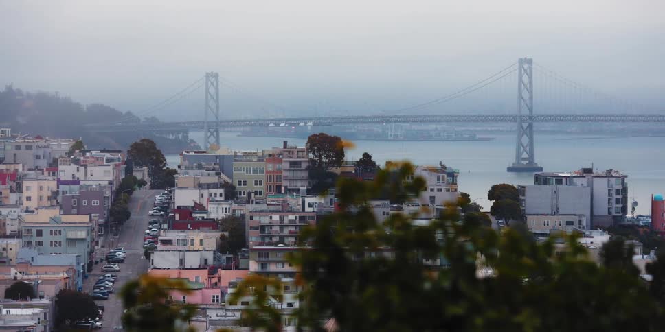 Cars line street passing apartment buildings as fog covers bridge area in the background