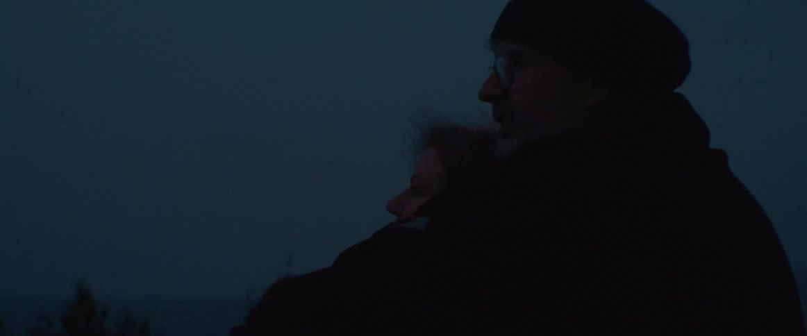 Young couple hugs near the ocean at night while a boat flashes a light on the distance