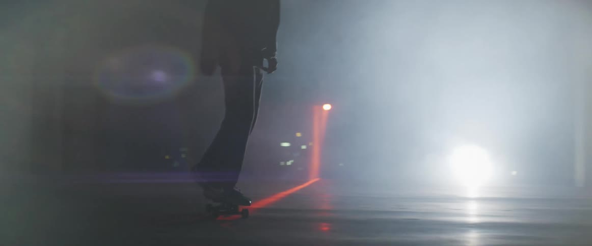 Man rides a skateboard on a dimly lit room filled with smoke near a red laser line on the floor