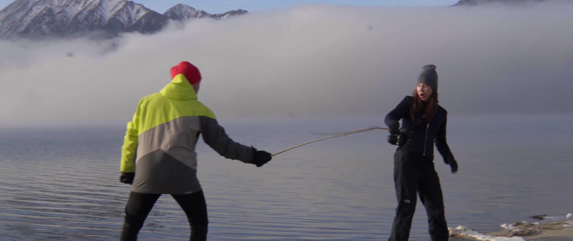 Two People Playfully Fight With Sticks by a Lake Sharing Laughter and Carefree Fun.