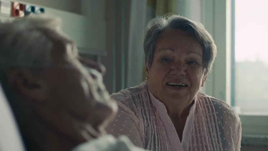 Old woman sitting next to her husband while hes laying down at the hospital bed